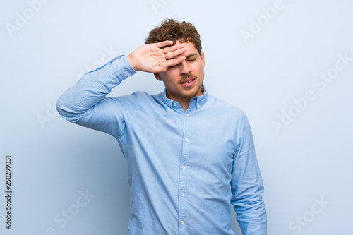 Blonde man over blue wall with tired and sick expression
