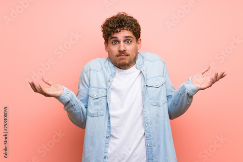 Blonde man over pink wall having doubts while raising hands and shoulders