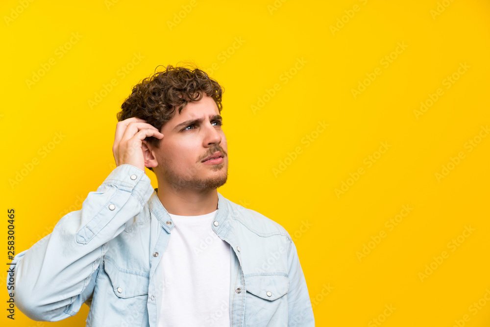 Blonde man over isolated yellow wall having doubts and with confuse face expression Blonde man over isolated yellow wall having doubts and with confuse face expression