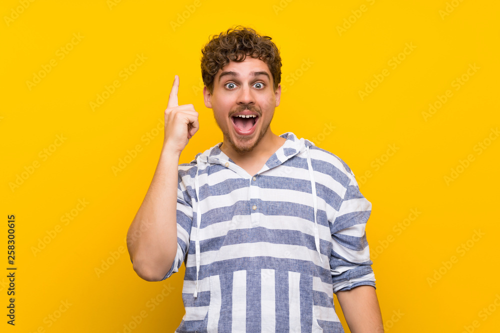 Blonde man over yellow wall intending to realizes the solution while lifting a finger up Blonde man over yellow wall intending to realizes the solution while lifting a finger up