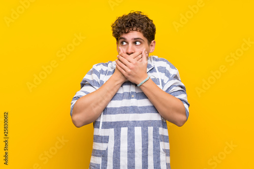 Blonde man over yellow wall covering mouth with hands