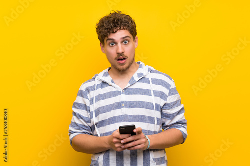 Blonde man over yellow wall surprised and sending a message