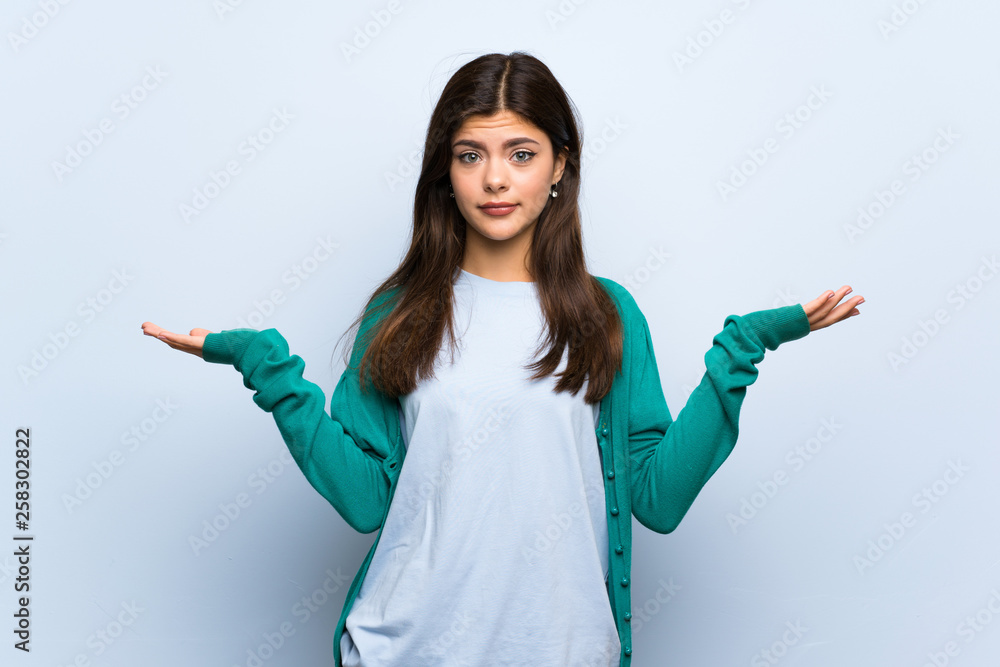 Teenager girl over blue wall having doubts while raising hands Teenager girl over blue wall having doubts while raising hands