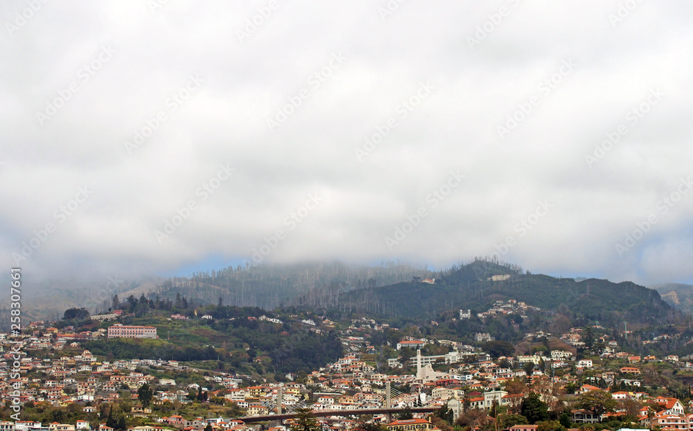 a panoramic view of the city of funchal with buildings and road bridge with mountains covered by white misty cloud