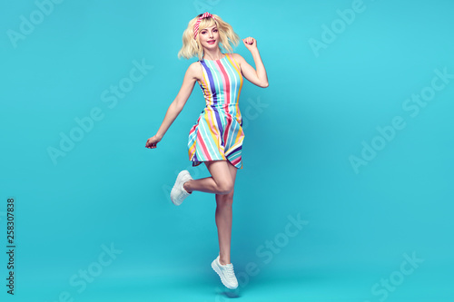 Fototapeta Naklejka Na Ścianę i Meble -  Fashionable shapely Young woman smiling on blue background. Excited blonde Girl Having Fun dance in Trendy summer dress, Stylish fashion sneakers, makeup. Happiness funny concept