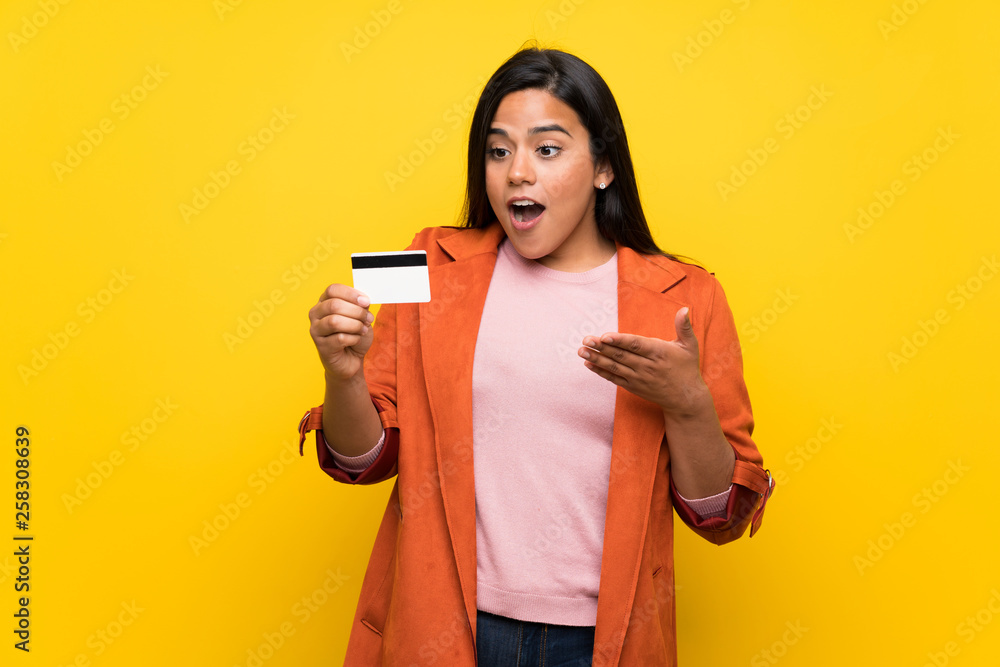 Young Colombian girl over yellow wall holding a credit card and surprised Young Colombian girl over yellow wall holding a credit card and surprised