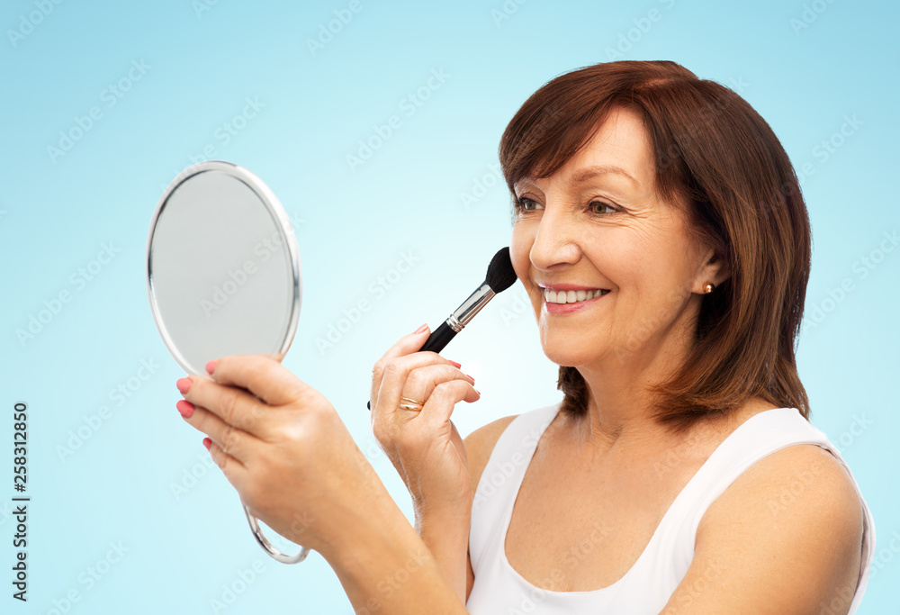 beauty, make up and old people concept - smiling senior woman with mirror applying blush by brush over blue background beauty, make up and old people concept - smiling senior woman with mirror applying blush by brush over blue background
