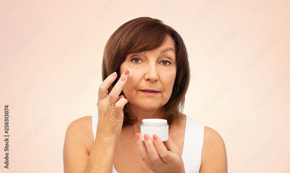 beauty, skincare and cosmetics concept - senior woman applying cream to her face over beige background beauty, skincare and cosmetics concept - senior woman applying cream to her face over beige background