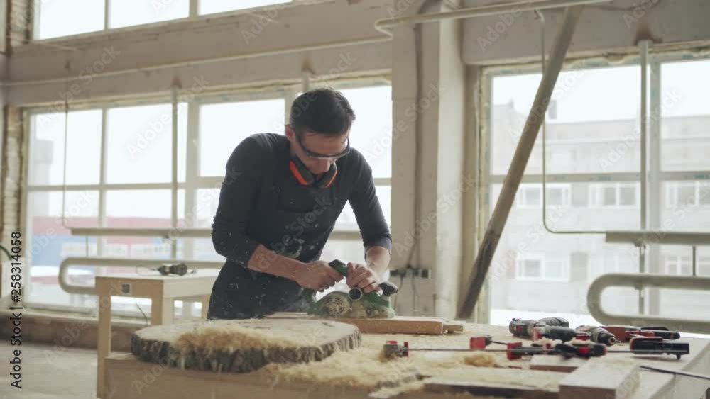 Panning shot of skilled carpenter sanding wooden plank with electric sanding machine, sawdust flying around Panning shot of skilled carpenter sanding wooden plank with electric sanding machine, sawdust flying around
