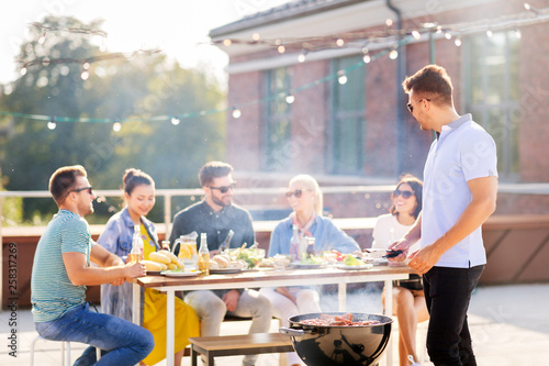 leisure and people concept - happy man grilling meat on bbq and friends at rooftop party