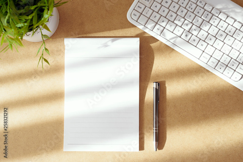Top view of workspace set up, on wooden table. Copy space on paper page from notepad, pen, white computer keyboard and green plant.