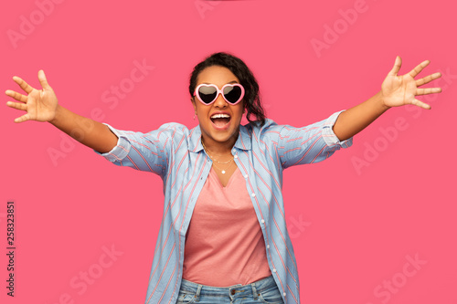 summer, valentine's day and emotion concept - happy african american young woman in heart-shaped sunglasses over pink background