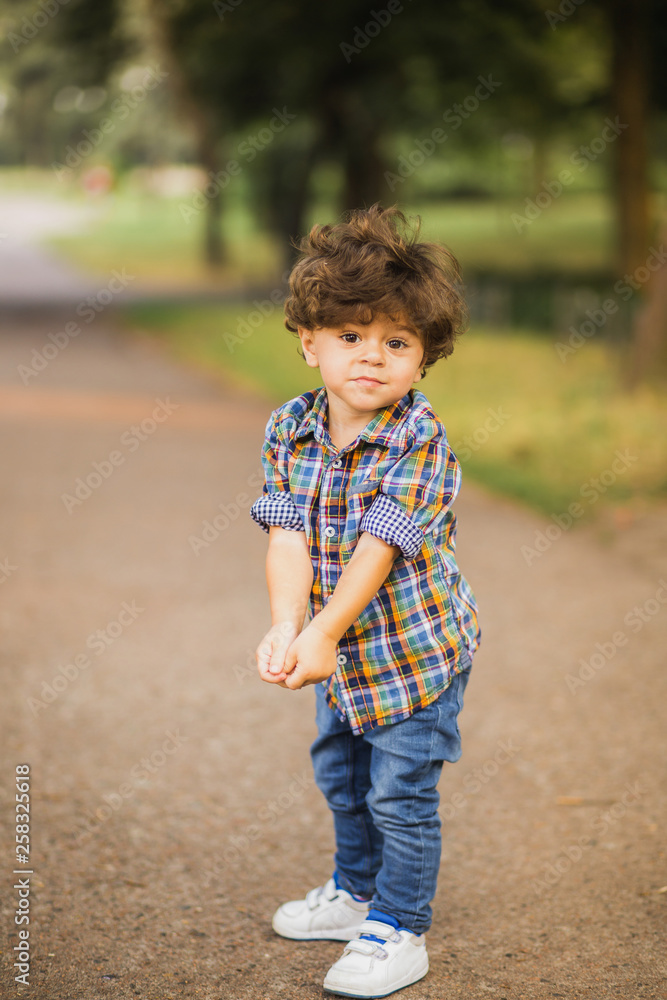 Portrait of handsome little toddler boy playing at summer city park. Calm child with beautiful curly brown hair posing for photo. Baby dressed in casual blue jeans and shirt. Vertical photography. Portrait of handsome little toddler boy playing at summer city park. Calm child with beautiful curly brown hair posing for photo. Baby dressed in casual blue jeans and shirt. Vertical photography.