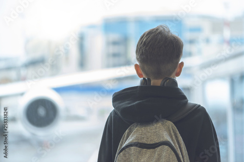A boy in headphones with a backpack stands near the window at the airport and looks at the plane. Departure hall