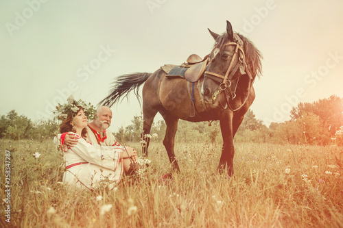 Mature man and woman in ethnic clothes are relaxing after a horse ride. Summer sunset on the background.