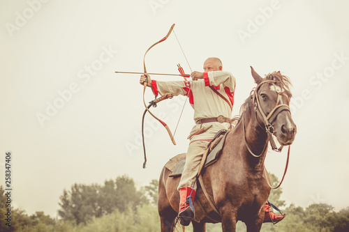 Man in ethnic clothing is riding a horse and aiming from the bow.