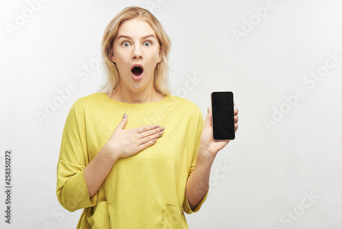 Shocked European blonde woman with mobile phone in hand, eyes bulging in surprise, emotions from what she sees on screen, keeps jaw dropped. Portrait of beautiful girl isolated in white studio