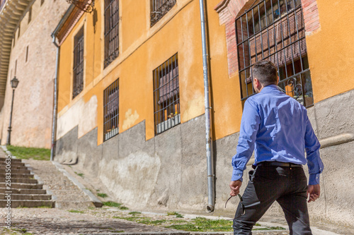 An elegant man in a blue shirt, walks through an old part of a city, on a sunny day © Óscar