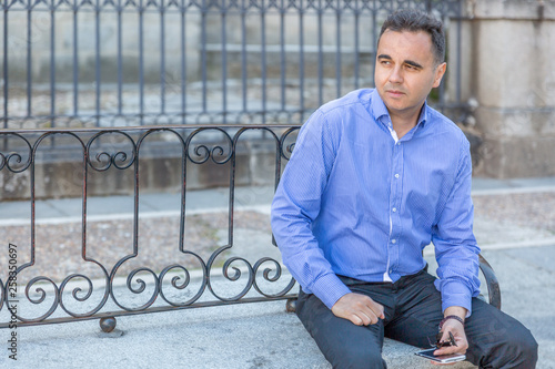 An elegant man in blue shirt waits sitting on a bench in the middle of a street