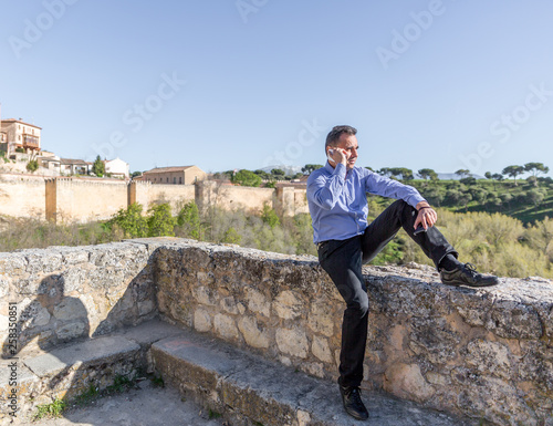 An elegant business man speaks by mobile phone in the outskirts of an old city, on a sunny day