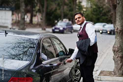 Stylish indian businessman in formal wear standing against black business car on street of city. Open door of car by key.