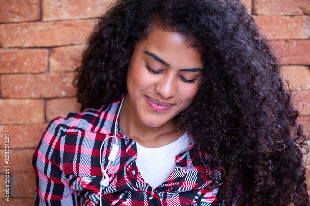 Happy student woman with afro hairstyle sitting with brick wall background using cell phone and smiling. Happy student woman with afro hairstyle sitting with brick wall background using cell phone and smiling.