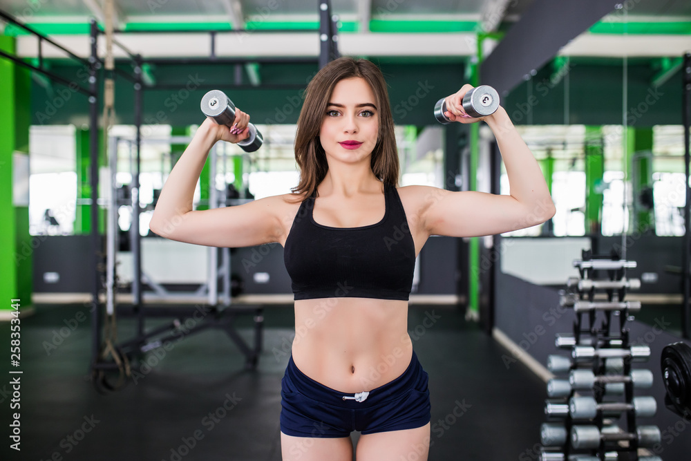 Fototapeta premium Young beautiful woman doing exercises with dumbbell in gym. Glad smiling girl is enjoying with her training process.