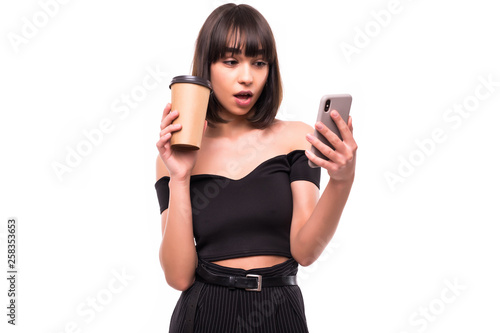 Portrait of a shocked surprised woman holding take away coffee cup and mobile phone while standing isolated over gray background