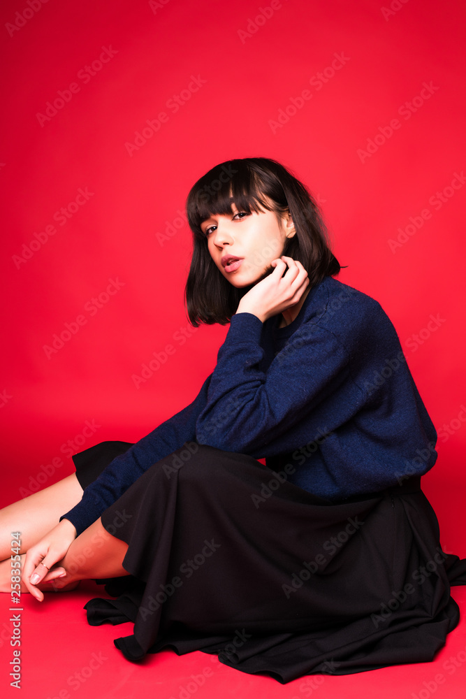 Laughing brunette sitting on the floor, pointing away and winks at the camera over red background Laughing brunette sitting on the floor, pointing away and winks at the camera over red background