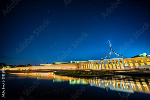 Facade of new parliament house in Canberra on capitol hill at sunset with bright illumination reflecting in blurred waters of pool.  South cross on sky  photo