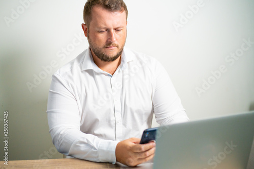 male in white shirt looks into the phone at the workplace sitting at a table near the computer