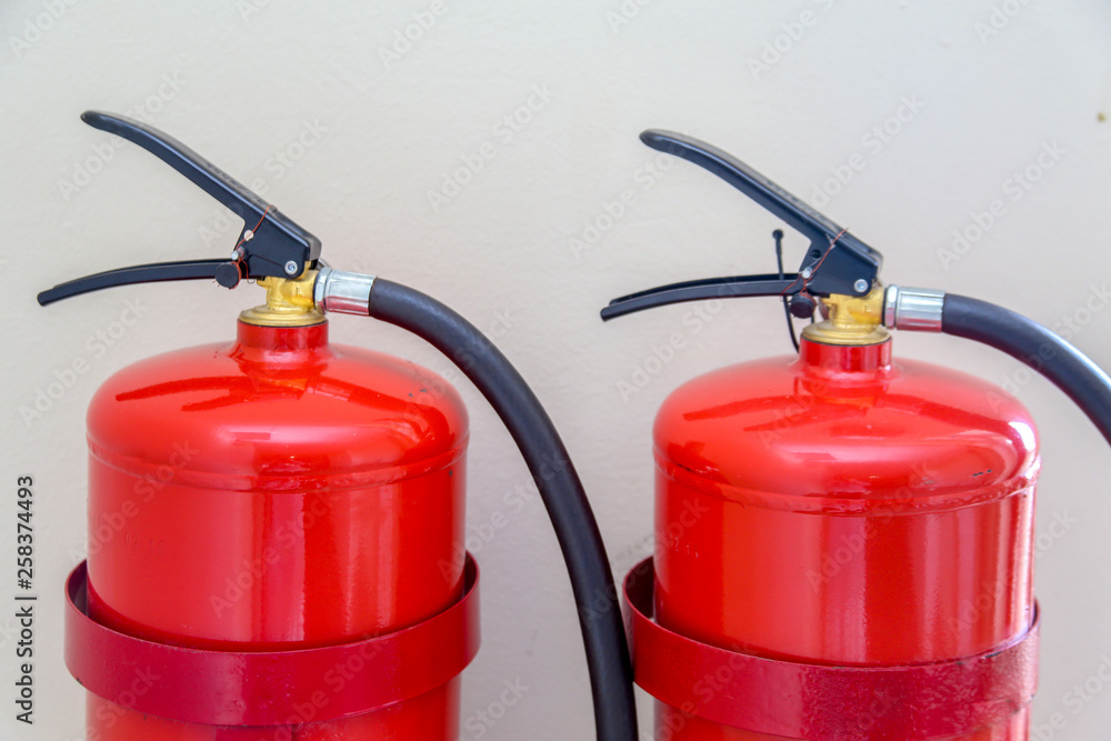 2 fire extinguishers hang on a light wall . 2 fire extinguishers hang on a light wall .