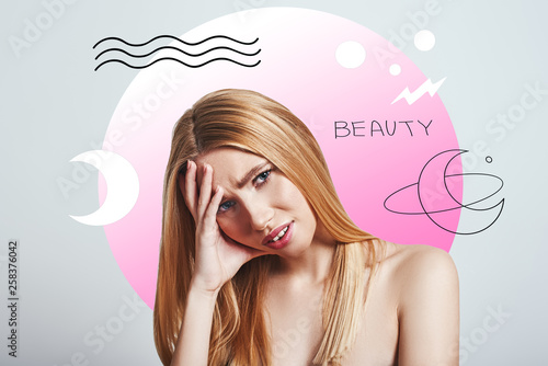 Not sure i am ok. Frustrated blonde woman making a face and keeping hand on head while standing against abstract pink circle and hand drawn illustrations.