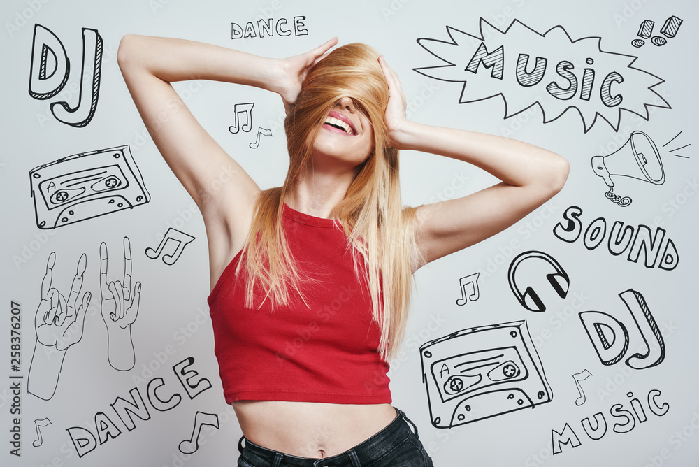 Fototapeta premium Feeling playful and happy. Slim woman in red tank top covering face with her hair and smiling while standing against grey background with music theme doodles.