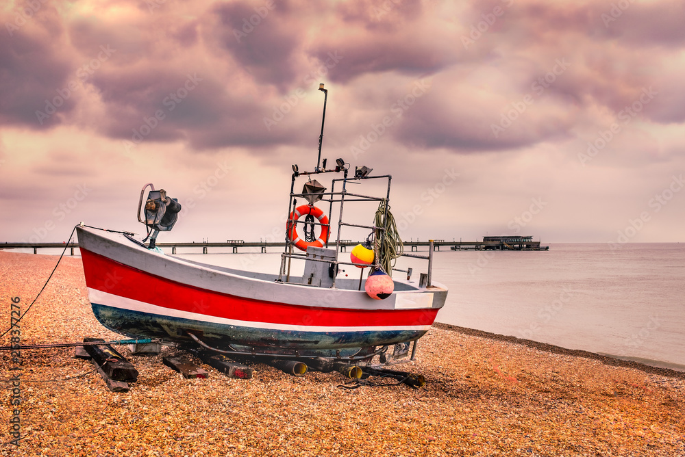 Red quaint, traditional fishing boat moored on the pebble shingle beach in Deal, Kent, UK with the pier in the background on a clouds winter day.