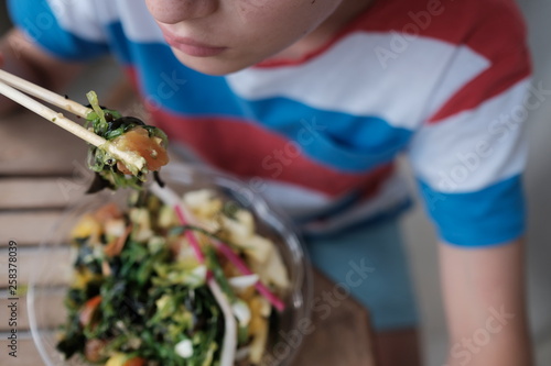 Young boy is eatinh healthy food poke bowl with chinese chopsticks. photo