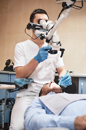 Care for your smile. Male dentist look at the patient using professional dental binocular microscope. Modern dental clinic
