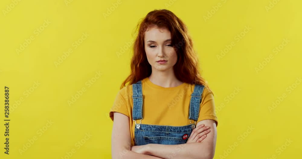 Portrait of the young Caucasian red haired girl looking at the camera and being not in the mood. Yellow background. Portrait of the young Caucasian red haired girl looking at the camera and being not in the mood. Yellow background.