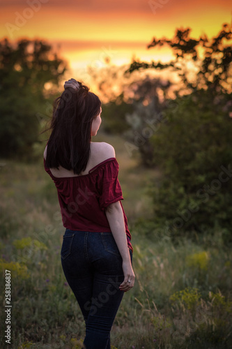 Girl in a red blouse and dark blue jeans stands with his back in nature. Sunset Sky Scarlett