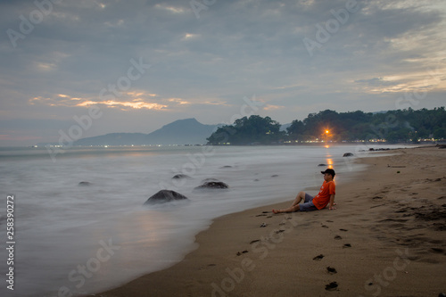 A man is sitting and enjoying the moment on Karang Hawu Beach, West Java, Indonesia with an orange sunset sky and blurry smooth waves photo