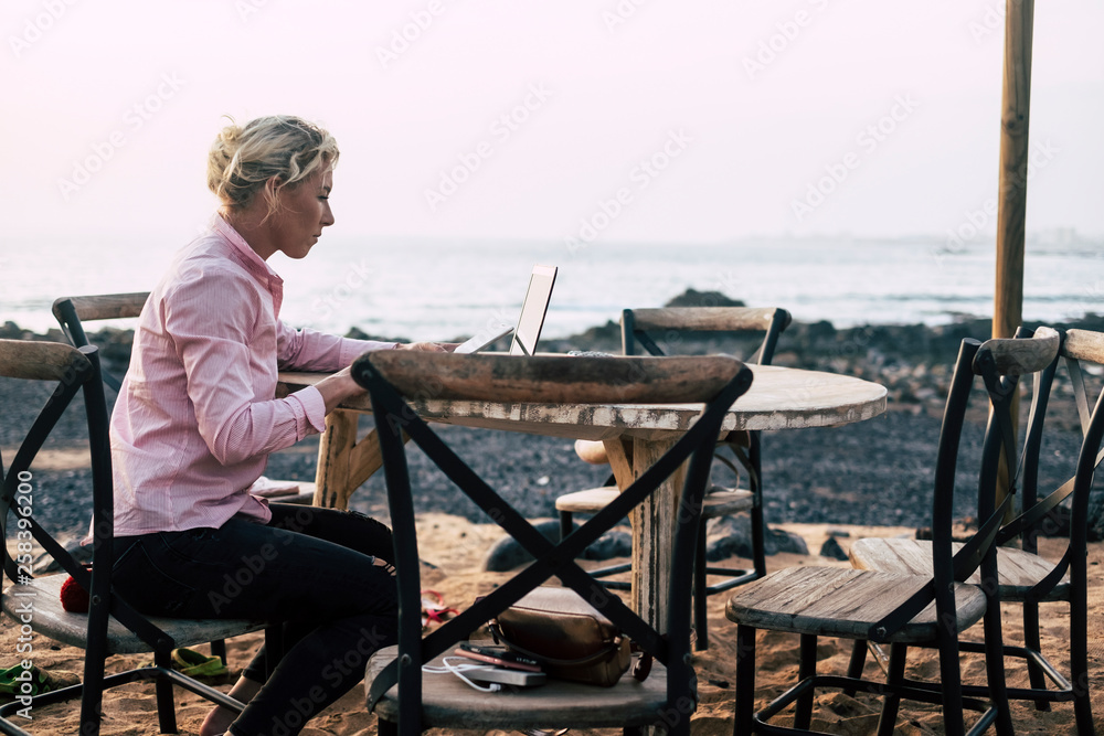 Young business woman sit down outdoor on a chair working on a wooden table with laptop - alternative office workstation near the ocean in totally freedom for modern people