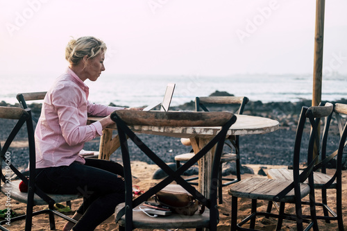 Young business woman sit down outdoor on a chair working on a wooden table with laptop - alternative office workstation near the ocean in totally freedom for modern people © simona