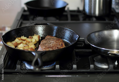 New York strip steak with potatoes frying in a cast iron pan on a natural gas stove top.
