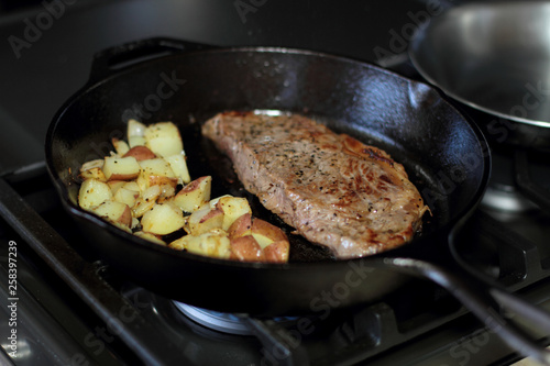 New York strip steak with potatoes frying in a cast iron pan on a natural gas stove top.