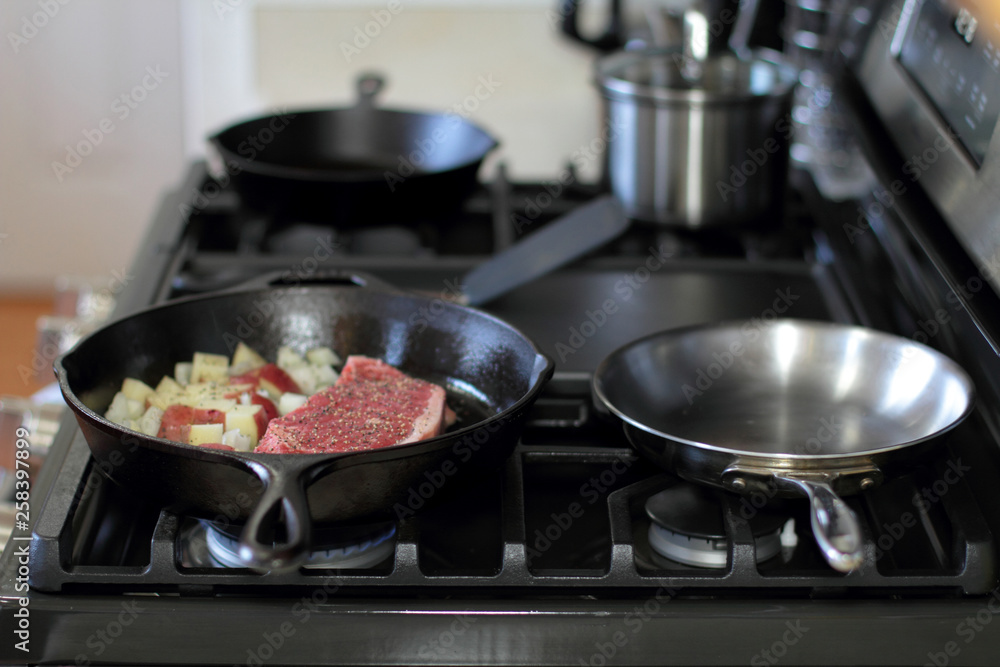 New York strip steak with potatoes frying in a cast iron pan on a natural gas stove top. New York strip steak with potatoes frying in a cast iron pan on a natural gas stove top.