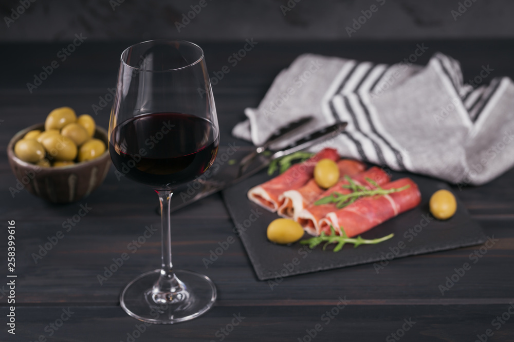 Glass of red wine with slices of cured ham or Spanish jamon serrano or Italian prosciutto crudo, green olives and arugula