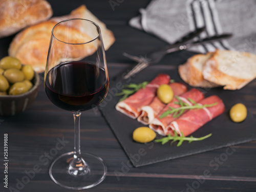 Glass of red wine with slices of cured ham or Spanish jamon serrano or Italian prosciutto crudo, bread, green olives and arugula