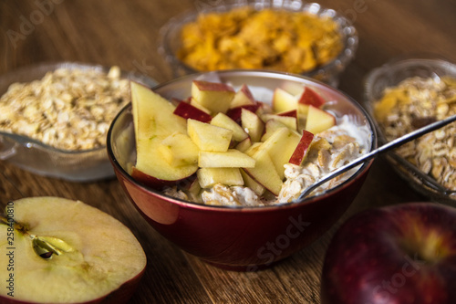 Muesli with dried fruit, milk and sliced red apple on wooden table. Barley flake, fresh organic red apple, and corn flakes in the background.