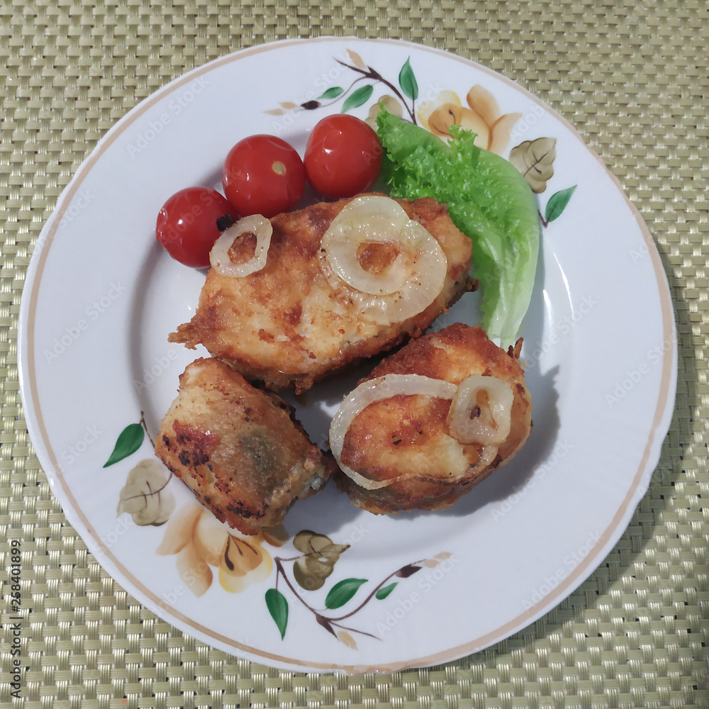 Three pieces of fried fish with onion rings in a plate on the table. Canned cherry tomatoes and greens are added. View from above