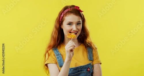 Wallpaper Mural Close up of the young Caucasian beautiful and stylish girl with red hair standing on the yellow wall background, holding a lollipop candy and smiling cheerfully. Portrait. Torontodigital.ca